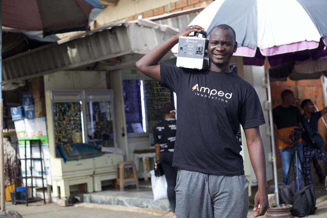 Ένα μέλος του προσωπικού της Amped Innovation κρατά μια από τις ηλιακές γεννήτριές της στον ώμο του—χαμογελώντας, φορώντας μαύρο μπλουζάκι Amped Innovation.